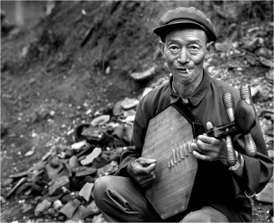#139-032a On a remote and rocky dirt road in the mountains of Sichuan, I came upon this easy-going truck driver plucking out a tune by the roadside for his own amusement. Drivers were a relatively privileged class in China. That explains his wristwatch. Travel in this era was restricted; one needed a letter from one’s production unit to buy a long-distance bus ticket or a train ticket. Air travel was even more restricted and favored people of high rank. The communists knew that freedom to travel freely around China had been essential for their organizing, fomenting and then leading their revolution to gain power. Party organizers had met secretly in hotels, which is why after they gained power they so closely monitor hotels in China. Guest lists must be turned in to the police every night. In 1979 the hotels even held guests’ passports until they departed just in case the police wanted to inspect or confiscate them. This driver is whiling away his time while waiting for an accident to be cleared or an argument over right-of-way to be settled. Either could take hours. That explains how I came to take the photo, i.e. my vehicle was also stalled in the same traffic jam. Travel would give someone like a truck driver opportunity for all kinds of graft and access to rare commodities. Shoes from factories often manage to leak out the back doors. A driver could buy up these shoes, put it in with his authorized load and take them to an area where shoes were scarce and thereby fetch a high price and so on. Giving a ride to someone unauthorized to travel could provide a small profit, enough to buy a wristwatch or a nice musical instrument.