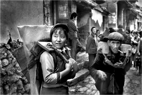 #175-034 These people carry their heavy loads long distances to reach the market in the mountain town of Lijiang, home to the Naxi minority. It may be a 2-3 day journey to return to their mountain villages. In 1987, it took me two twelve-hour days of hard driving on bumpy dirt roads to get to Lijiang. Deeply remote, there were no hotels and only one government hostel. However, its very isolation preserved its fabulously interesting culture and spectacular scenic vistas. During WWII, the Flying Tigers had a landing field here. Today, it is a major tourist attraction.
